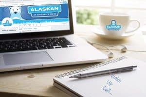 Desk view of laptop showing Alaskan website, coffee cup with Alaskan logo, and note book and pen with note "wake up call..."