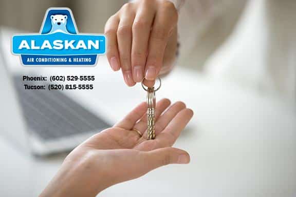 One hand giving another open hand a set of keys, laptop open in background, with Alaskan Air Conditioning and Heating logo and contact phone numbers in top left corner