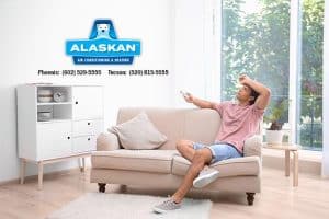 man sitting on couch, hand to forehead, holding remote, Alaskan Air Conditioning and Heating logo and contact phone numbers in top left quadrant