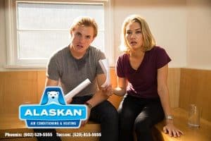 Man and woman sitting together, each holding papers, looking shocked / angry at camera Alaskan Air Conditioning and Heating logo and contact phone numbers in bottom left corner