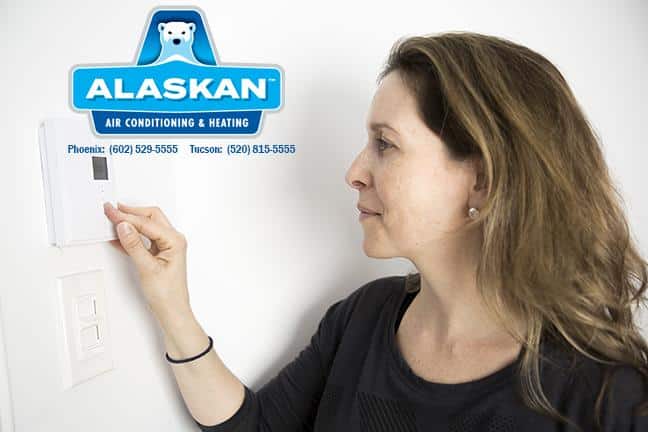 Woman adjusting thermostat on wall with Alaskan Air Conditioning and Heating logo and contact phone numbers in top left quadrant